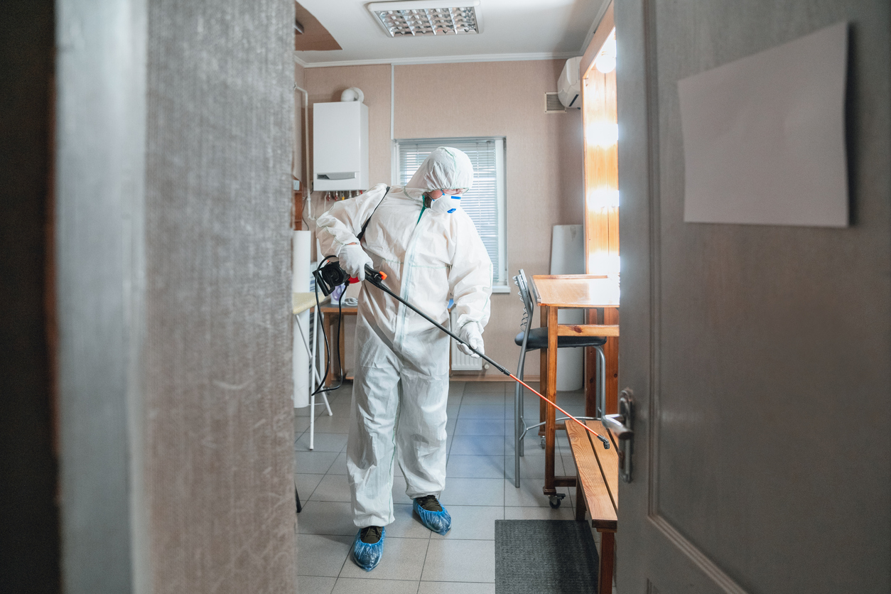 A technician in a Tyvek suit holds a sprayer as they walk through a commercial space, spraying to sterilize.