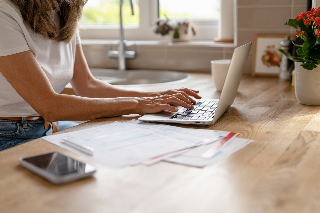 A woman at a kitchen counter working on a laptop with insurance paperwork and documents spread out in front of her.